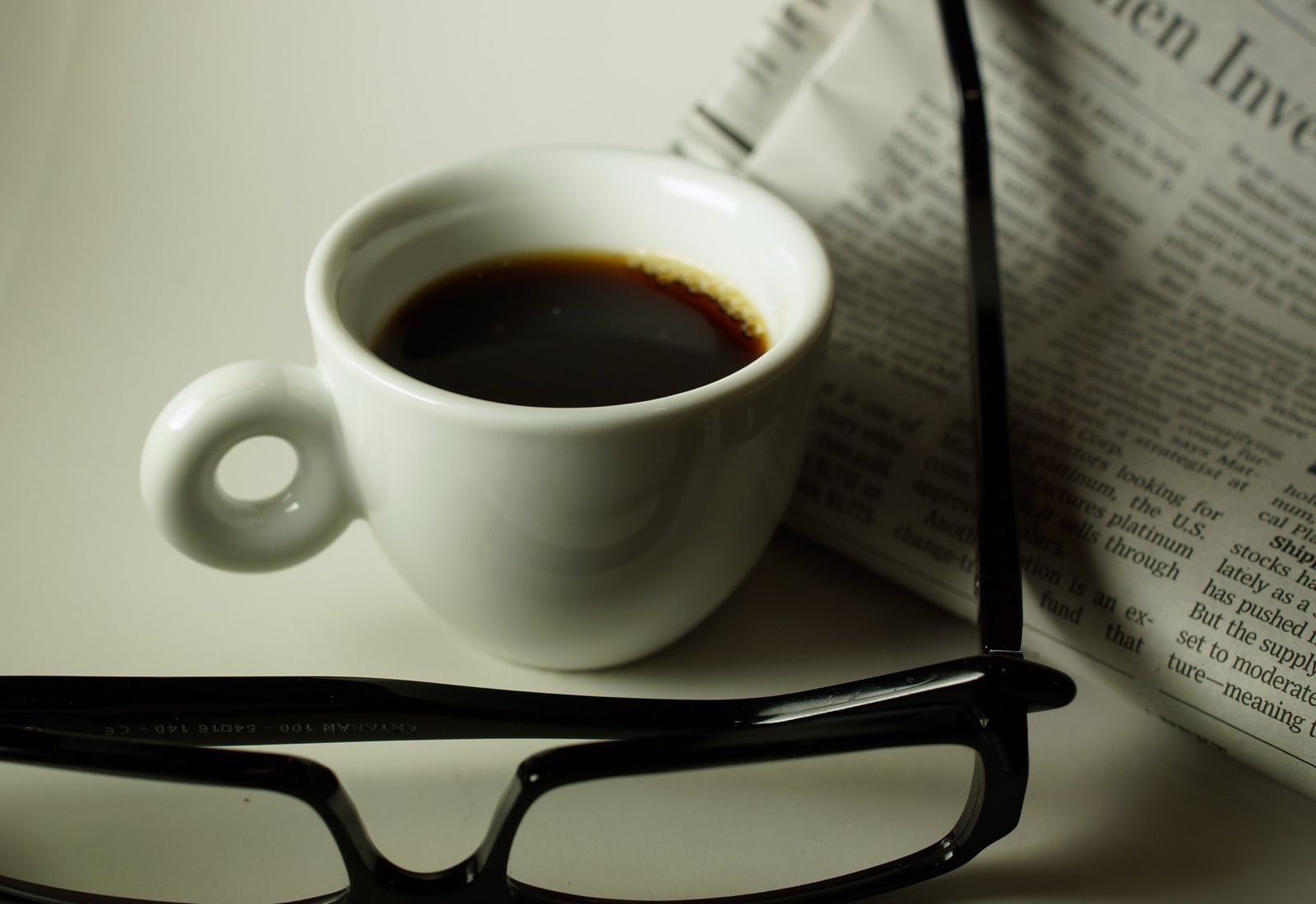 A cup of black coffee beside a folded newspaper and a pair of glasses